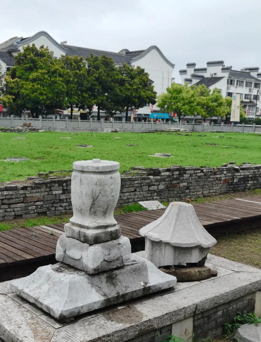 白色台座上两件石质构件：左侧盖罐形、右侧屋脊形，背景为树木与民居